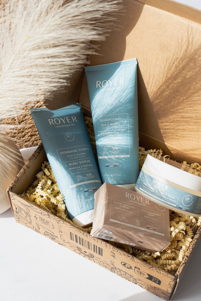 Un ensemble de produits cosmétiques bio de la marque Royer, comprenant un shampoing, un gommage pour le corps, un baume et un savon, disposés dans une boîte en carton remplie de papier de soie.